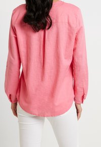 Camicia a maniche lunghe in lino rosa con orlo arrotondato, piega sul retro e polsini abbottonati, abbinata a pantaloni bianchi.