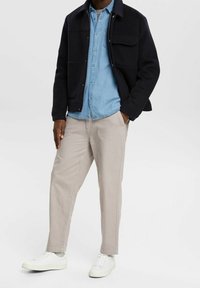 Chaqueta de mezcla de lana azul marino sobre una camisa de mezclilla azul claro, combinada con pantalones beige y zapatillas blancas; con botones delanteros y bolsillos laterales.