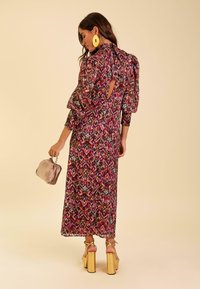Vestido maxi con un vibrante estampado floral multicolor, tejido transparente, mangas abullonadas, espalda descubierta, combinado con un bolso de mano con detalles dorados y tacones de plataforma.