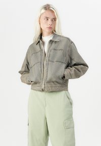 Verkorte denimjas in gedempt grijs met zichtbare stiksels, voorkruiken en een ritsluiting. Draag het over een lichte trui en groene broek.
