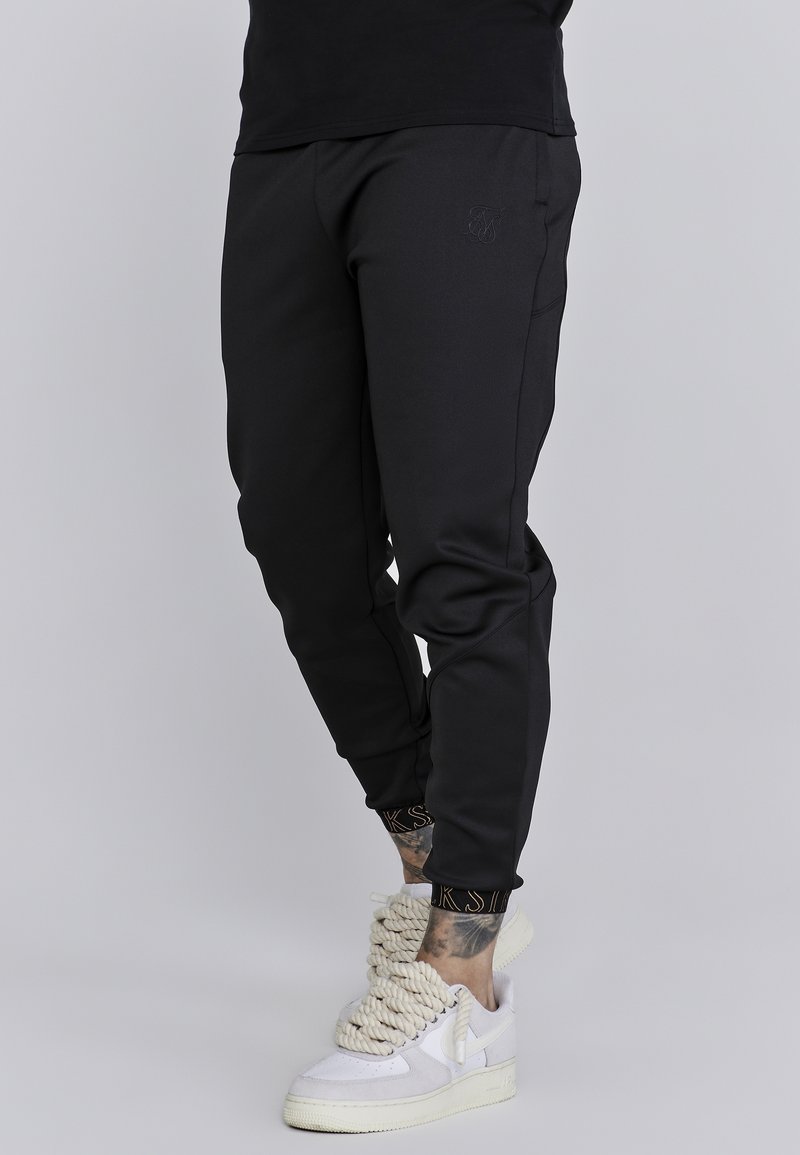 Zwarte joggers met een slanke pasvorm, voorzien van een logo op het bovenbeen en geribbelde manchetten. Gecombineerd met witte sneakers met unieke veters.
