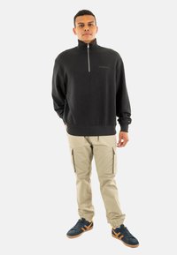 Maglione zip-up nero con texture a coste, pantaloni cargo beige e scarpe blu con dettagli arancioni. Il modello sta in piedi dritto contro uno sfondo grigio.