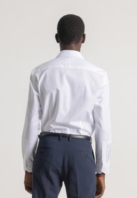 Hombre vestido con una camisa de vestir de manga larga blanca metida dentro de unos pantalones azul marino con un cinturón negro, visto desde atrás frente a un fondo liso.