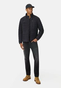 Sort quiltet jakke med høj krave, lagdelt over en sort skjorte, parret med mørke jeans og brune støvler. Modellen har en matchende kasket på.