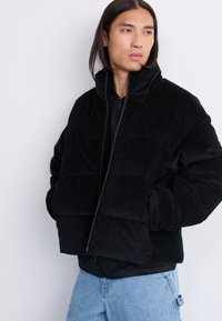 Giacca puffer in corduroy nero con collo alto, chiusura a zip e due tasche frontali. Il design presenta sezioni trapuntate e una vestibilità rilassata.