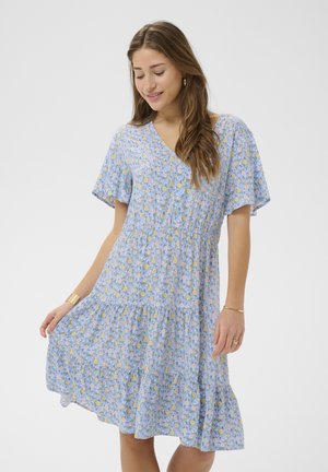 Jonge vrouw die een blauwe, knielange jurk met bloemenprint en korte mouwen draagt, de rok vasthoudt en glimlacht met gesloten ogen tegen een effen achtergrond.