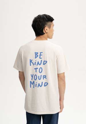 Mann mit dem Rücken zur Kamera, trägt ein beiges T-Shirt mit blauem Text "BE KIND TO YOUR MIND" und dunkle Hose vor einem schlichten weißen Hintergrund.