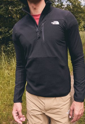 Mężczyzna w czarnym polarze The North Face i beżowych szortach stoi na zewnątrz na trawiastym szlaku, ramiona swobodnie opuszczone wzdłuż ciała, głowa częściowo poza kadrem.