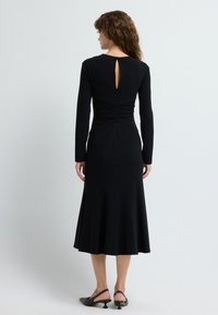 Schwarzes Langarmkleid mit tailliertem Oberteil, schwingendem Rock, Raffungen am Rücken und einem Schlüsselloch-Ausschnitt am Hals.