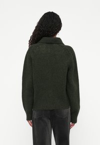 Pulover verde tricotat cu guler înalt, având o croială lejeră, tiv cu nervuri și o textură tricotată deschisă pe spate. Asortat cu pantaloni închiși la culoare.