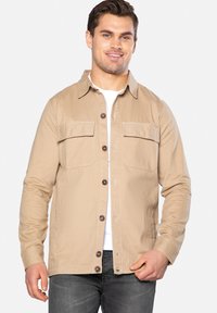 Beige jakke lavet af bomuld med to brystlommer, knaplukning og brune knapper. Båret over en hvid skjorte, parret med grå jeans.