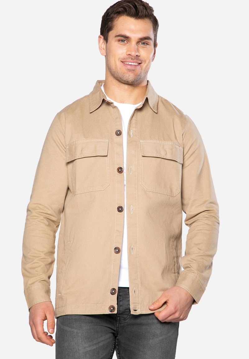 Beige jakke lavet af bomuld med to brystlommer, knaplukning og brune knapper. Båret over en hvid skjorte, parret med grå jeans.
