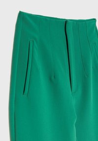 Pantaloni verdi su misura con vita alta, due tasche laterali e pieghe nette. Il tessuto ha una texture morbida e una vestibilità strutturata.