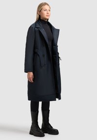 Marineblauer langer Mantel aus glänzendem Stoff, mit doppelter Knopfleiste, großen Revers, seitlichen Taschen und einem taillierten Gürtel. Schwarze Schnürstiefel.