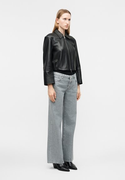 Ενήλικη γυναίκα που στέκεται, φορώντας μαύρο δερμάτινο μπουφάν με φερμουάρ, ανοιχτόχρωμα γκρι τζιν με φαρδιά μπατζάκια και μαύρες μπότες με μυτερή μύτη, σε απλό φόντο.