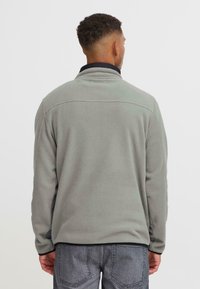Mann iført lys grå fleecejakke og mørkegrå jeans, sett fra bak på en ensfarget lys bakgrunn.