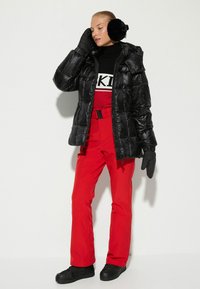 Chaqueta de plumas negra con capucha, pantalones rojos de tiro alto con cinturón, guantes negros y orejeras de piel sintética. Zapatos negros.