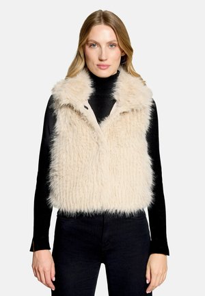 Woman with long blonde hair wearing a beige fluffy vest over a black long-sleeve top and black pants, facing forward with neutral expression.