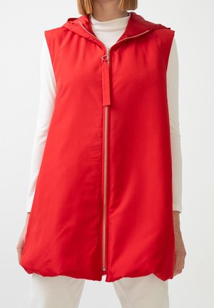 Ärmellose rote gesteppte Weste mit Kapuze und Frontreißverschluss, getragen über einem weißen Langarmshirt und weißen Hosen.