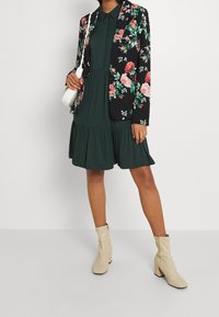 Mujer vistiendo un vestido de capas verde oscuro, una chaqueta negra con estampado floral, botas de tobillo crema y sosteniendo un bolso blanco contra un fondo sencillo.