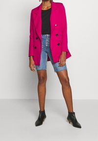 Fuchsiový dvouřadý blazer s klopy s výřezy, kombinovaný s džínovými krátkými kalhotami s vysokým pasem. Černé kotníkové boty dokreslují vzhled.