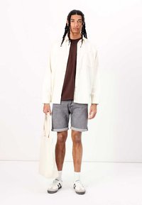 Langærmet creamfarvet skjorte, brun T-shirt, grå rullede shorts, lys grå sneakers, hvide sokker og en tote bag i naturstof.