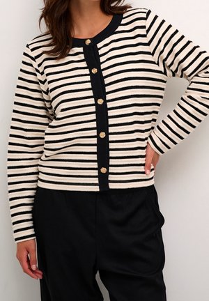Femme portant un cardigan rayé horizontal noir et blanc avec des boutons dorés et un pantalon noir, la main posée sur la hanche.