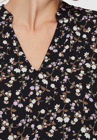 Schwarzer Stoff mit floralem Muster, das weiße und lavendelfarbene Blumen zeigt, mit einem V-Ausschnitt-Design und subtilen texturalen Details des Drucks.