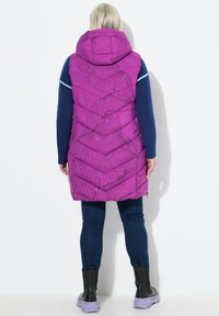 Padded fialová vesta s kapucí, zdobená abstraktními černými liniemi a nadýchaným designem. Nositelná přes modrý top a kombinovaná s tmavými džíny a botami.