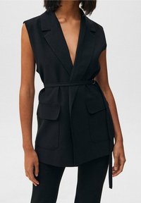 Schwarzer ärmelloser Blazer mit tiefem V-Ausschnitt, zwei Fronttaschen und einem Gürtel in der Taille. Glatte Textur mit tailliertem Schnitt.