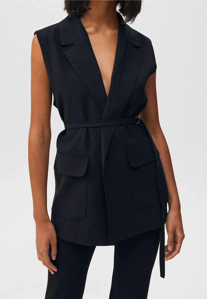 Schwarzer ärmelloser Blazer mit tiefem V-Ausschnitt, zwei Fronttaschen und einem Gürtel in der Taille. Glatte Textur mit tailliertem Schnitt.