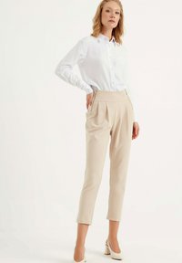 Beige op maat gemaakte broek met plooien, ritsen op de zakken, gecombineerd met een wit overhemd met knopen en witte blokhakken.