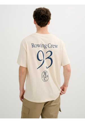 Hombre con camiseta beige "Rowing Crew 93" y pantalones caqui, mostrado de espaldas, de pie frente a un fondo claro liso.