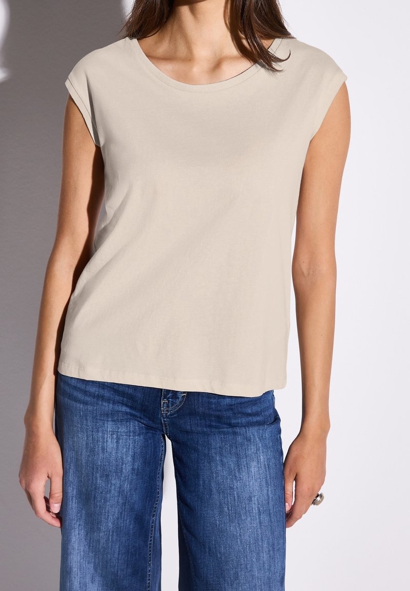 T-shirt beige in cotone con maniche a cappuccio, scollo rotondo e una leggera caduta. Abbinato a jeans a gamba larga blu, che evidenziano un look casual.