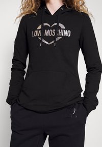 Sweat à capuche noir avec un logo "LOVE MOSCHINO" argenté métallique en forme de cœur. Dispose d'une poche kangourou et d'une capuche à cordon de serrage ajustable.