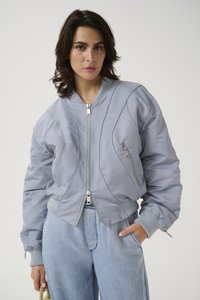 Giacca bomber grigia con silhouette aderente, texture trapuntata, zip visibili e polsini a costine a contrasto. Abbinata a pantaloni in denim chiaro.