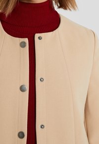 Abrigo beige con botones de presión frontales sobre un suéter de cuello alto acanalado en rojo intenso. Presenta una textura suave, forma entallada y diseño minimalista.