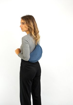 Sac circulaire bleu en matériau doux, porté dans le dos. Surface lisse avec un minimum de détails, associé à un haut à manches longues gris et un pantalon noir.