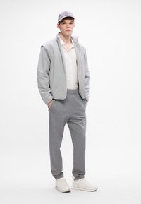 Chaqueta acolchada gris claro con interior texturizado, camiseta blanca, pantalones de chándal grises y zapatillas blancas. Gorra gris con ala y logo.