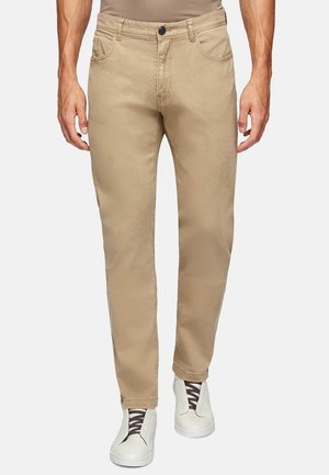 Pantalones de algodón beige con un corte recto, diseño de cinco bolsillos, trabillas para cinturón y cierre de botón, combinados con zapatos blancos tipo slip-on.