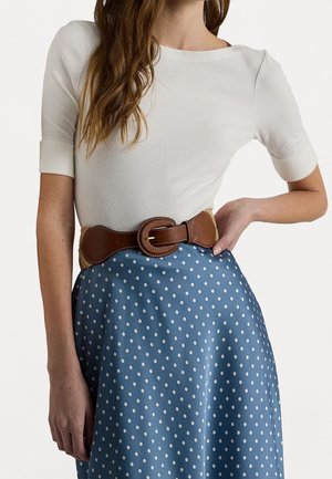 Bruine leren riem met stikdetails gedragen over een blauwe polkadot rok. Het bovenstuk is een strakke witte stof met een ronde halslijn.