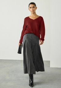 Nainen kävelee sisätiloissa burgundinpunaisessa pitkähihhaisessa paidassa, harmaissa leveälahkeisissa housuissa, mustissa teräväkärkisissä saappaissa ja pitää kädessään pientä mustaa clutch-laukkua.