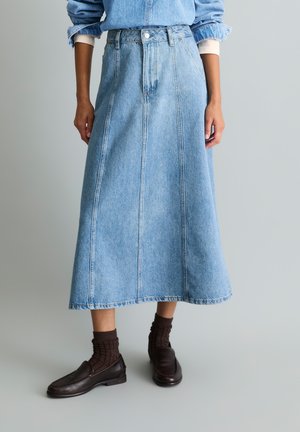 Femme portant une jupe midi en denim bleu clair avec coutures visibles, associée à des chaussettes marron foncé et des mocassins noirs sur un fond uni.