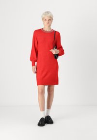 Robe en tricot rouge avec un col rond et des manches bouffantes longues. Le modèle porte des chaussures noires et porte un petit sac noir. Texture lisse.
