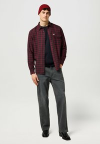 Camicia di flanella a quadri rossi e neri, t-shirt nera sotto, jeans grigi, stivali neri e un berretto di lana rosso, in piedi su uno sfondo neutro.