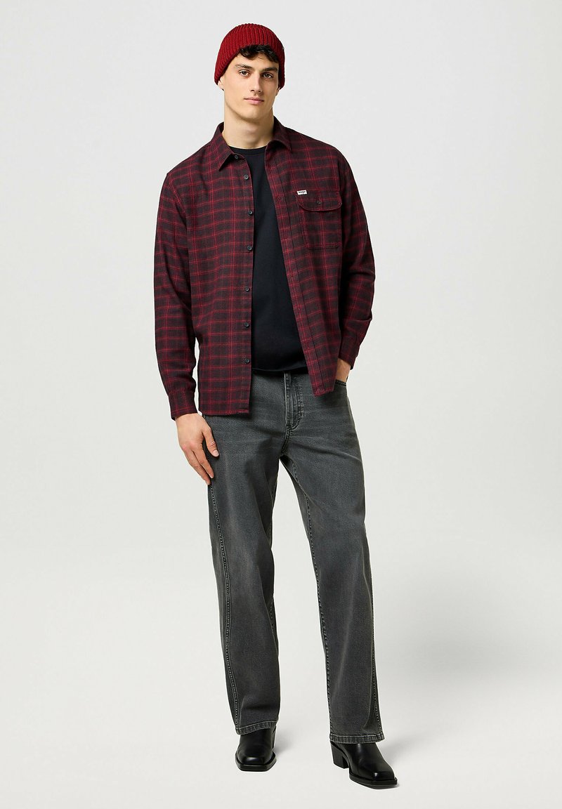 Camicia di flanella a quadri rossi e neri, t-shirt nera sotto, jeans grigi, stivali neri e un berretto di lana rosso, in piedi su uno sfondo neutro.