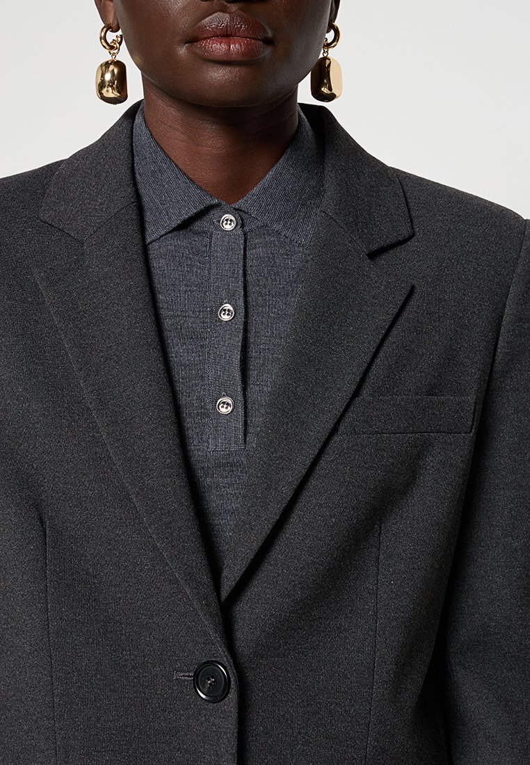 Blazer grigio con un design su misura, caratterizzato da rever a punta, chiusura con un solo bottone e una tasca sul petto sopra una camicia colletto grigio scuro.