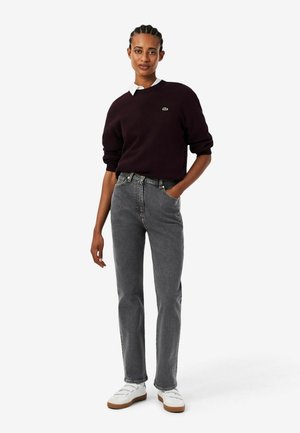 Vrouw draagt een donkerpaarse trui, wit overhemd met kraag, grijze jeans en witte sneakers, staand met één hand in de zak tegen een witte achtergrond.