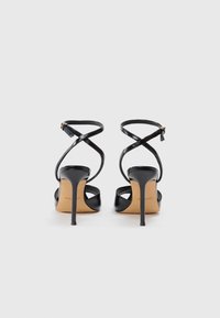 Sandalias de tacón alto negras con tiras, con un acabado brillante, que presentan tiras cruzadas en el tobillo y un delgado tacón de aguja. Suela beige con detalle del logo.