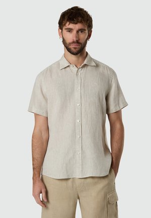 Hombre con camisa de manga corta abotonada color beige claro y pantalones cargo beige, de pie contra un fondo claro y sencillo.